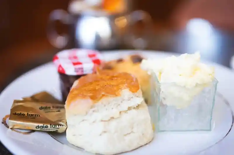 A plate featuring two scones, a small container of clotted cream, a jar of jam with a red and white checkered lid, and two butter packets.