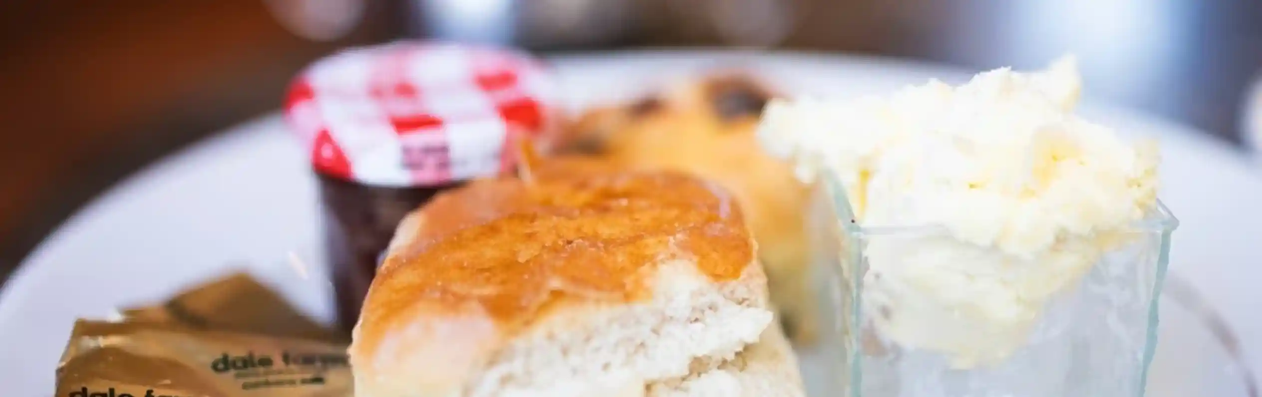 A plate featuring two scones, a small container of clotted cream, a jar of jam with a red and white checkered lid, and two butter packets.