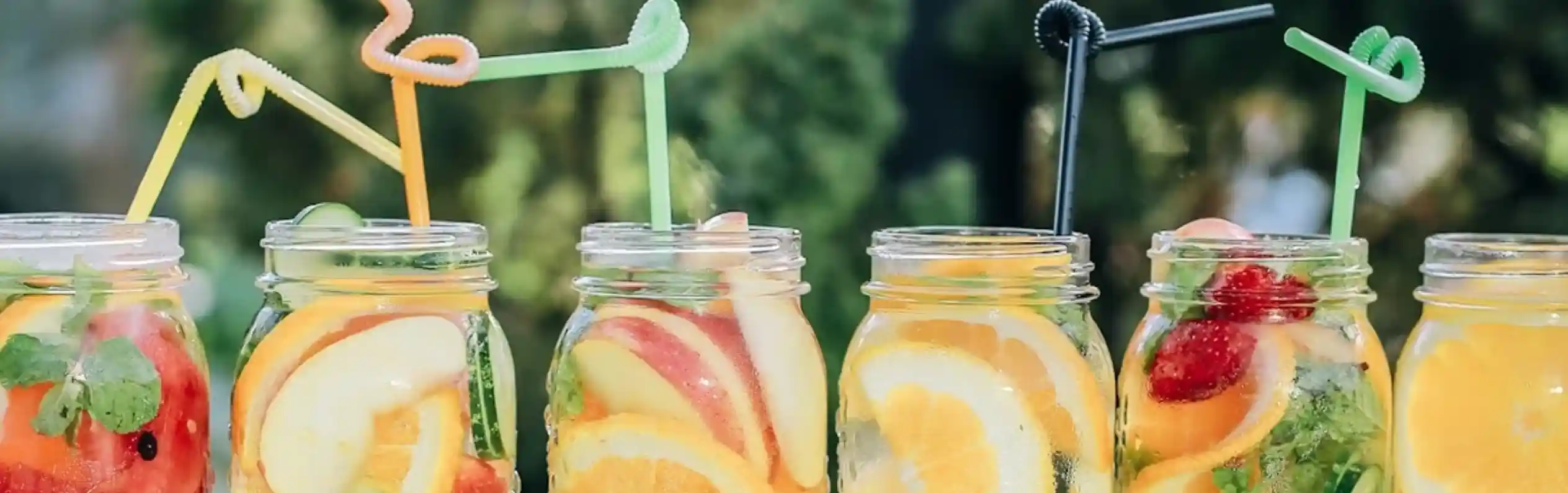Colorful fruit-infused drinks in mason jars lined up on a ledge, each with a unique combination of citrus fruits and fresh herbs, adorned with playful straws.