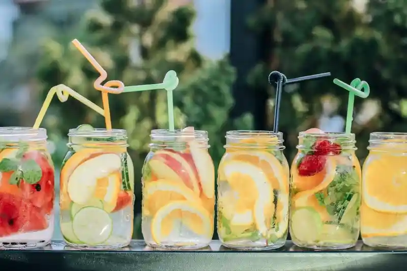 Colorful fruit-infused drinks in mason jars lined up on a ledge, each with a unique combination of citrus fruits and fresh herbs, adorned with playful straws.