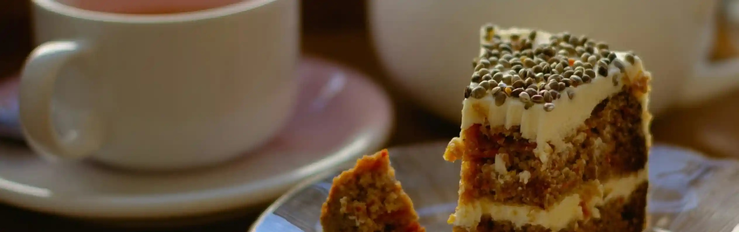 A slice of layered cake with cream frosting on a plate, next to a cup of tea and a teapot. The cake features visible layers with a crumbly texture and decorative toppings.