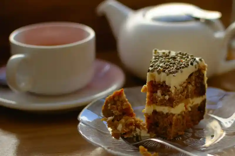 A slice of layered cake with cream frosting on a plate, next to a cup of tea and a teapot. The cake features visible layers with a crumbly texture and decorative toppings.