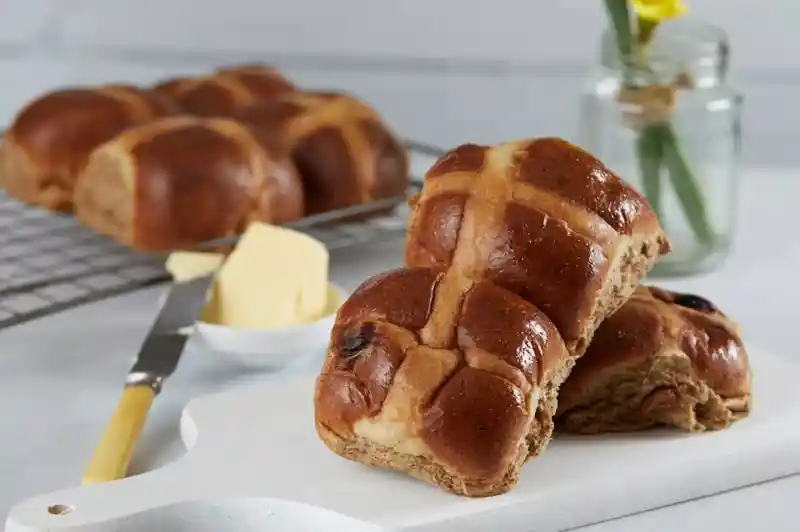 Freshly baked hot cross buns on a white cutting board, with a pat of butter and a butter knife beside them. A small jar containing a yellow flower is in the background.