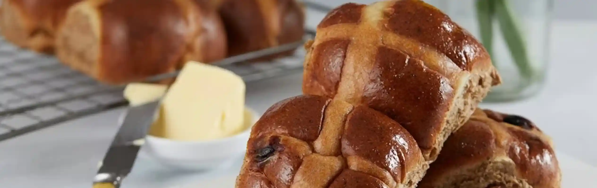 Freshly baked hot cross buns on a white cutting board, with a pat of butter and a butter knife beside them. A small jar containing a yellow flower is in the background.