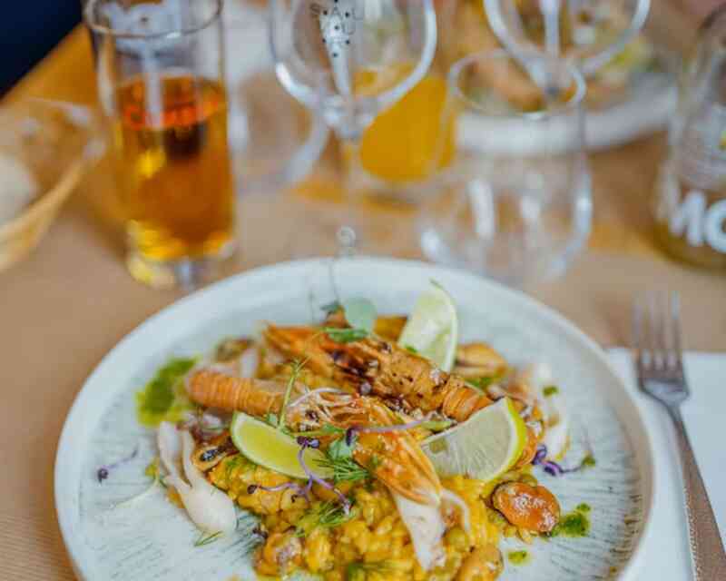 A colorful plate featuring a seafood dish with shrimp, grilled chicken, and garnishes of lime, herbs, and colorful microgreens, served on a decorative white plate. Behind the dish are glasses and a beer, accompanied by a casual dining setting.