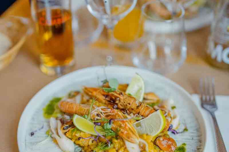 A colorful plate featuring a seafood dish with shrimp, grilled chicken, and garnishes of lime, herbs, and colorful microgreens, served on a decorative white plate. Behind the dish are glasses and a beer, accompanied by a casual dining setting.