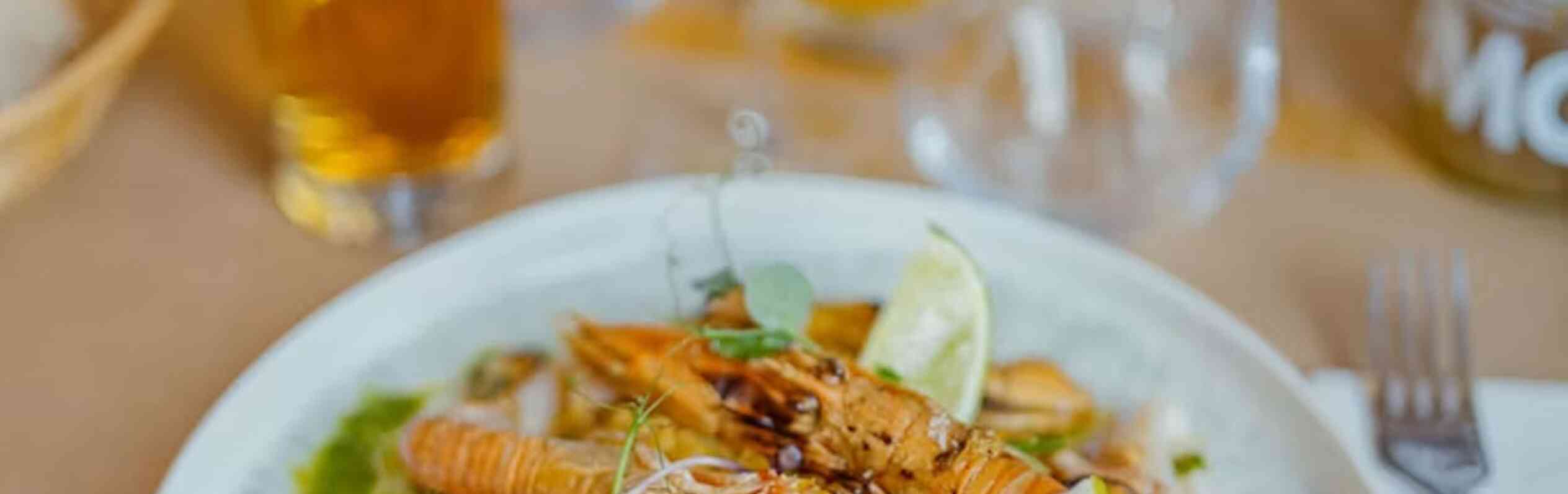 A colorful plate featuring a seafood dish with shrimp, grilled chicken, and garnishes of lime, herbs, and colorful microgreens, served on a decorative white plate. Behind the dish are glasses and a beer, accompanied by a casual dining setting.