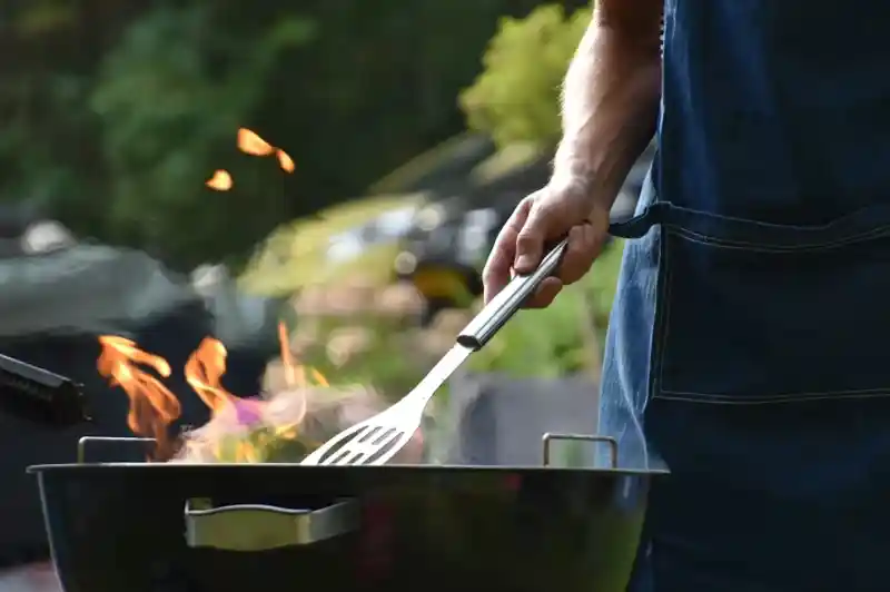 A person wearing a denim apron uses a spatula to flip food on a barbecue grill, with flames rising from the grill. A lush garden is blurred in the background.