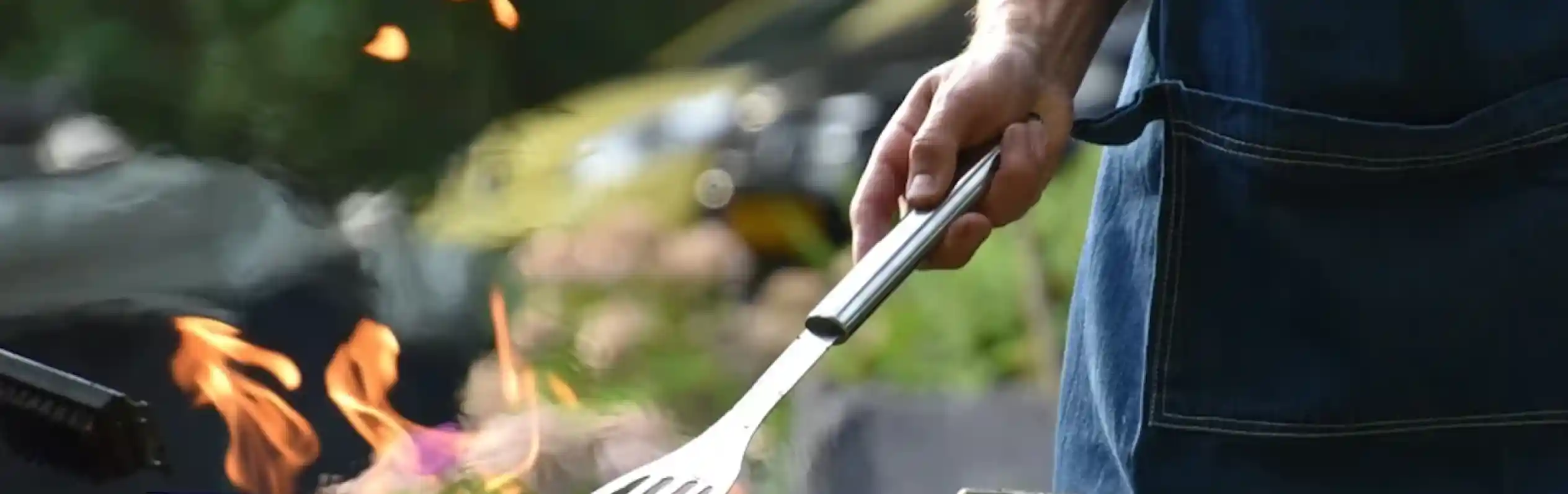 A person wearing a denim apron uses a spatula to flip food on a barbecue grill, with flames rising from the grill. A lush garden is blurred in the background.