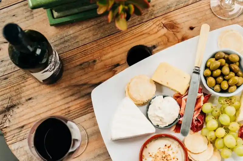 A wooden table displays a cheese and charcuterie platter featuring various cheeses, olives, grapes, and crackers. A glass of red wine sits beside the platter, along with a bottle of wine and a small potted plant in the background.