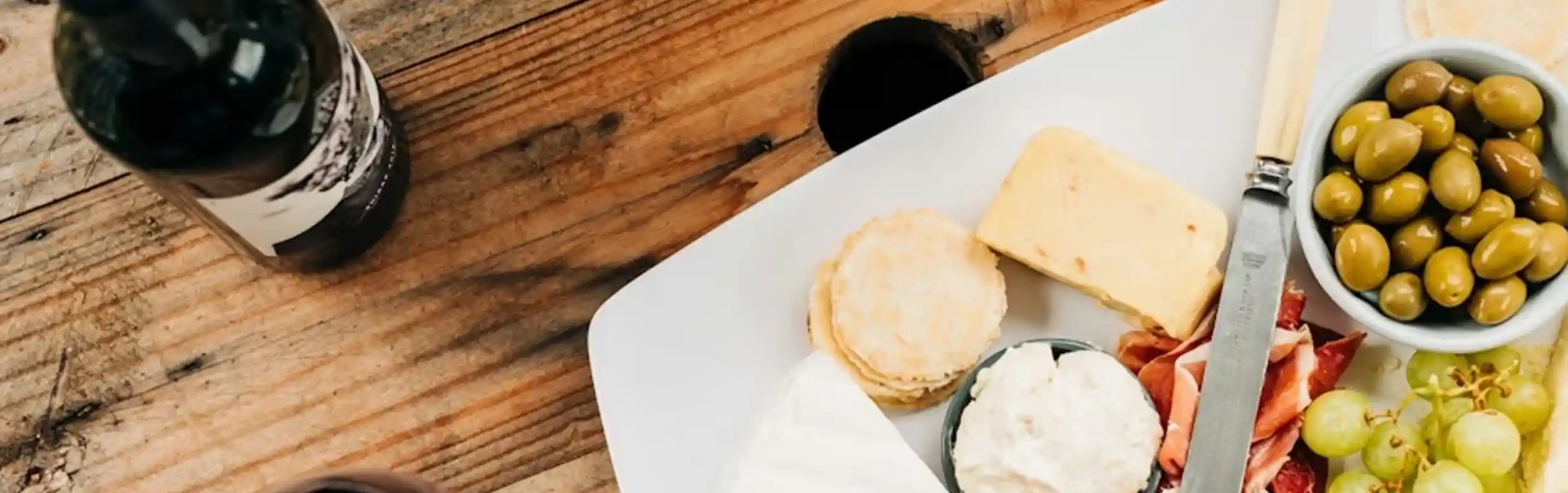 A wooden table displays a cheese and charcuterie platter featuring various cheeses, olives, grapes, and crackers. A glass of red wine sits beside the platter, along with a bottle of wine and a small potted plant in the background.