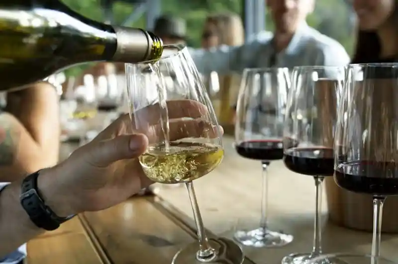 A hand pours white wine into a elegantly shaped glass, placed beside several glasses of red wine on a wooden table. In the background, a group of people enjoys a social gathering.