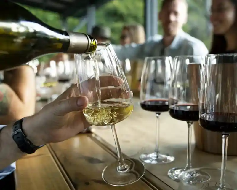 A hand pours white wine into a elegantly shaped glass, placed beside several glasses of red wine on a wooden table. In the background, a group of people enjoys a social gathering.