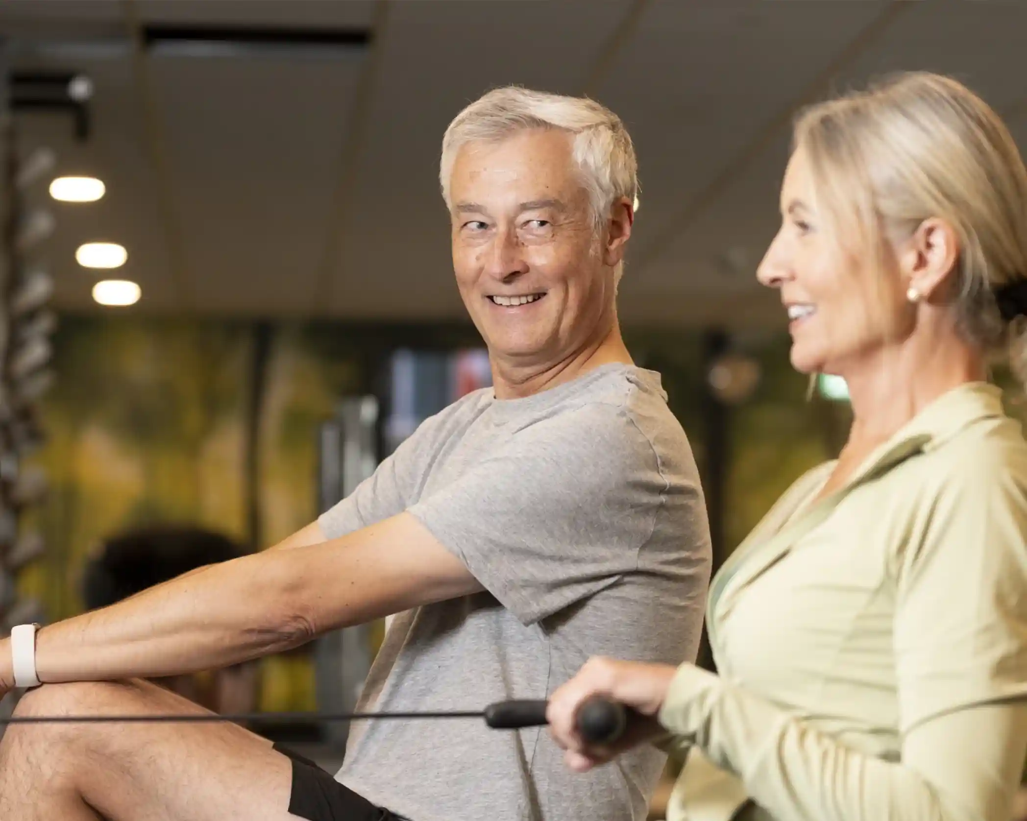 A smiling older man and a woman exercise together in a gym, using rowing machines. The man looks at the camera, while the woman smiles ahead, both appearing engaged and focused on their workout. The background shows gym equipment and a vibrant mural.
