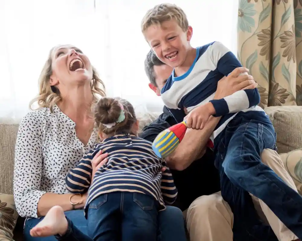 A family enjoying a playful moment together on a couch. A woman laughs while holding a young girl, and a boy playfully runs around, smiling and holding a toy. The atmosphere is joyful and lively.