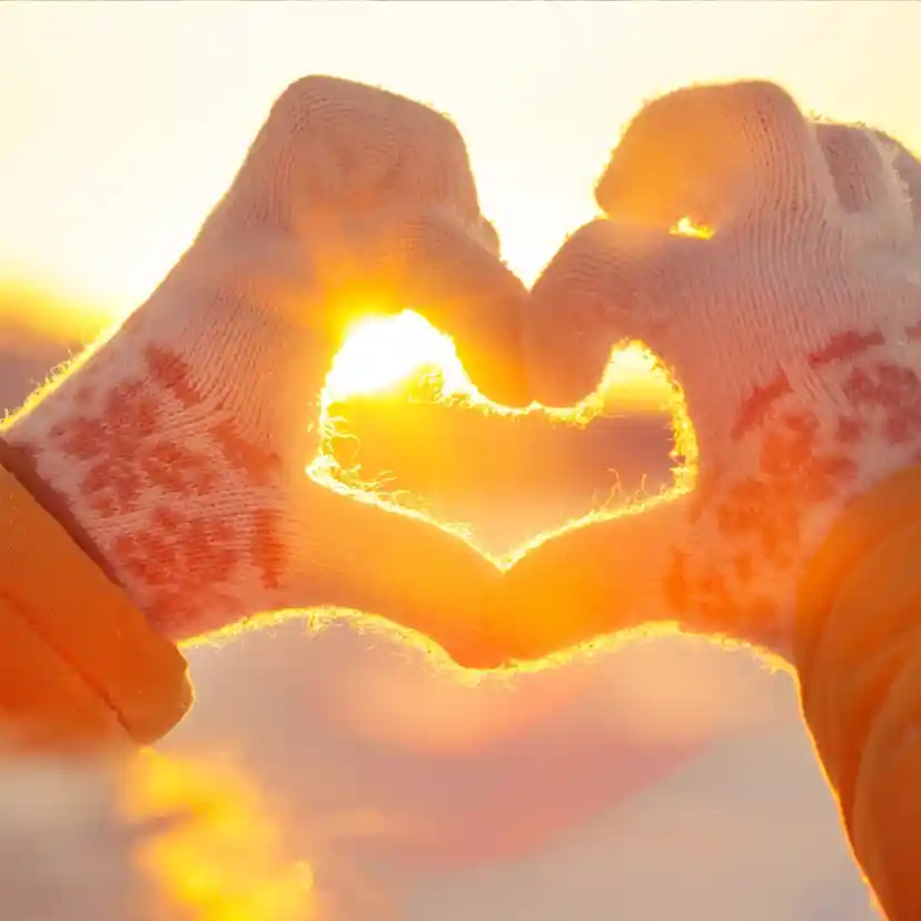 Hands wearing knitted gloves form a heart shape against a bright sunset, with sunlight glowing through the fingers. The background is softly blurred, creating a warm and romantic atmosphere.