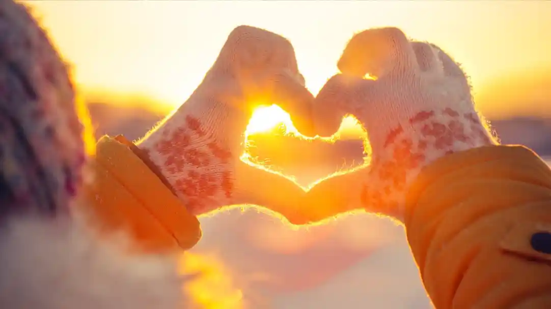 Hands wearing knitted gloves form a heart shape against a bright sunset, with sunlight glowing through the fingers. The background is softly blurred, creating a warm and romantic atmosphere.