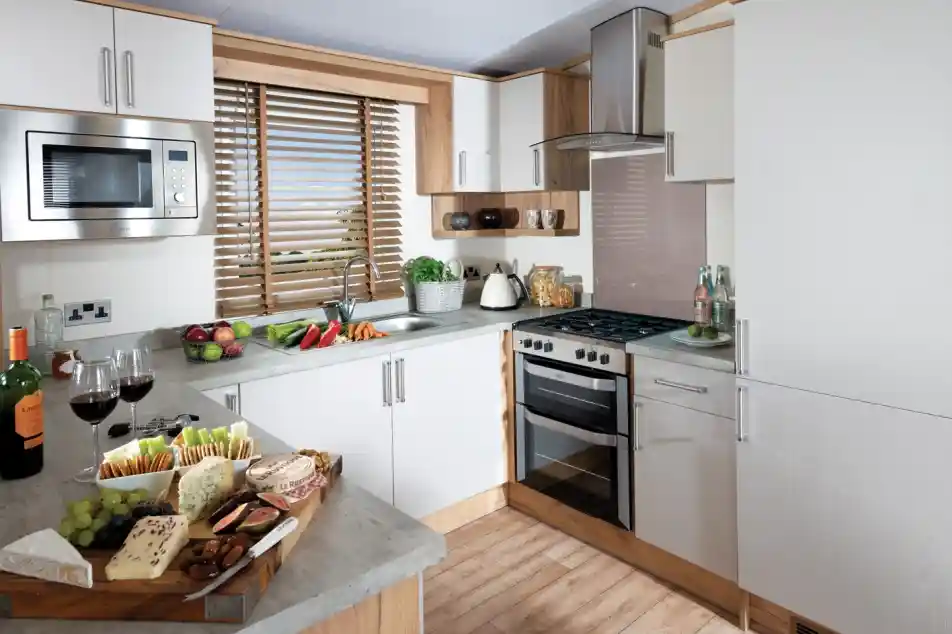 Modern kitchen with white cabinets, wooden accents, and grey countertops. A platter with assorted cheeses, grapes, and two glasses of red wine sits on the counter. A window with wooden blinds lets light in, creating a warm, inviting atmosphere.