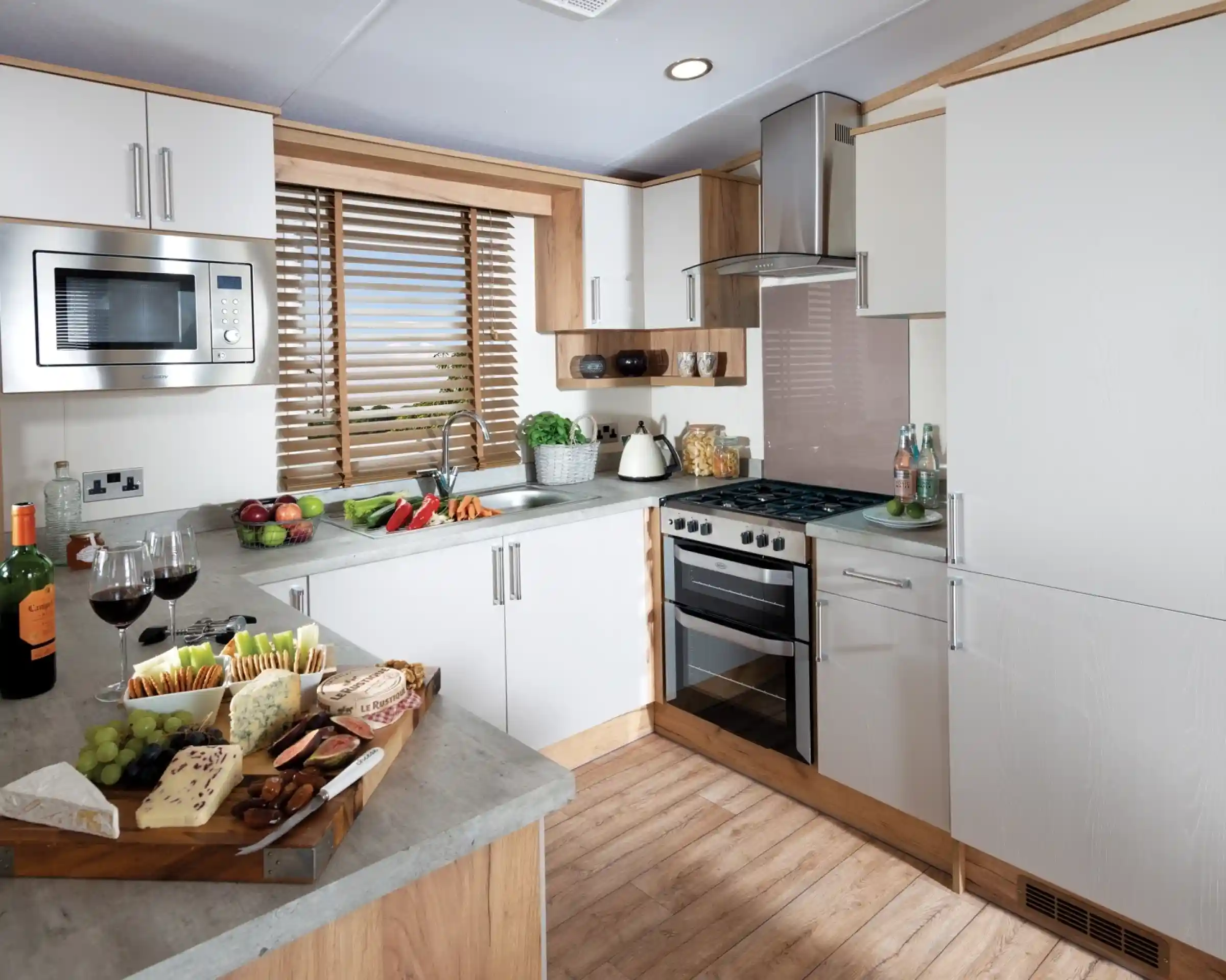 Modern kitchen with white cabinets, wooden accents, and grey countertops. A platter with assorted cheeses, grapes, and two glasses of red wine sits on the counter. A window with wooden blinds lets light in, creating a warm, inviting atmosphere.