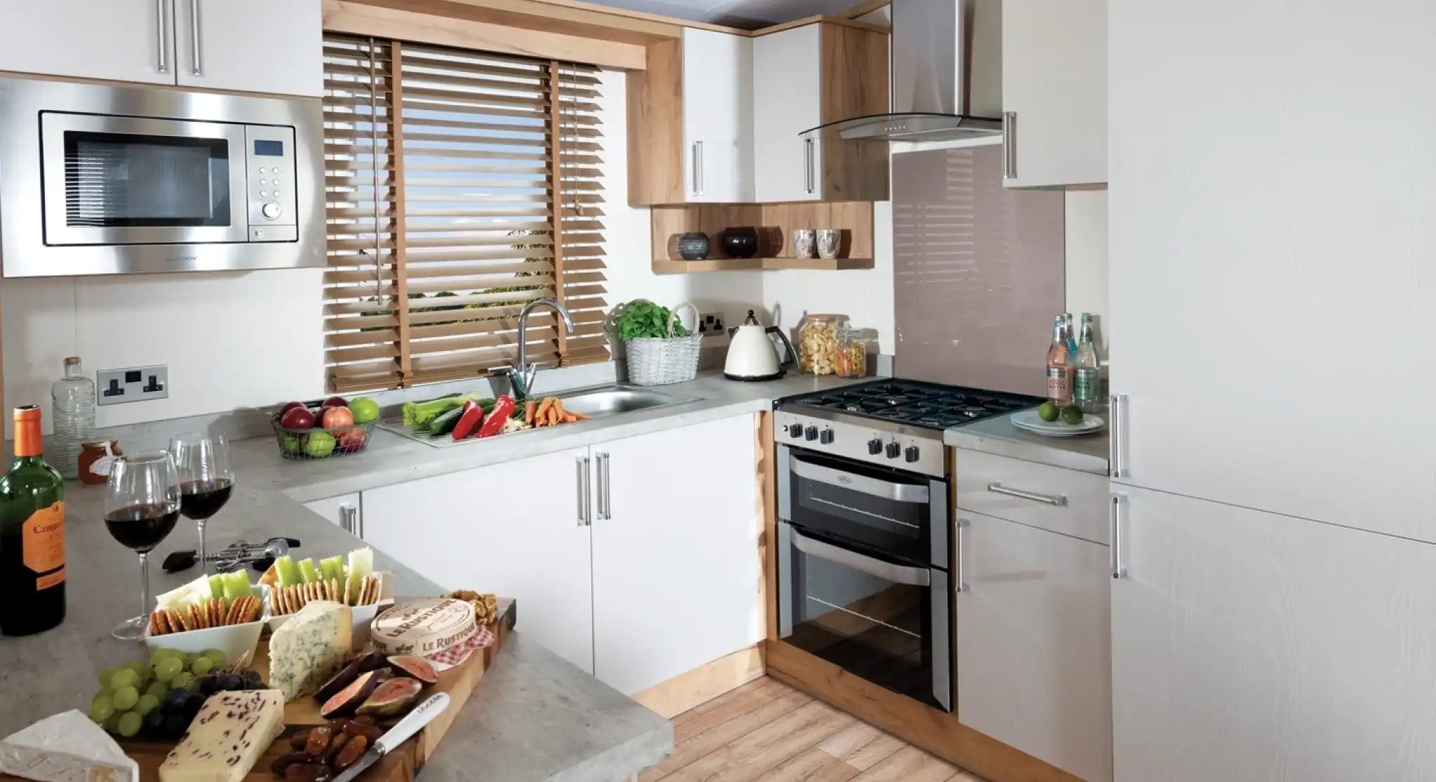 Modern kitchen with white cabinets, wooden accents, and grey countertops. A platter with assorted cheeses, grapes, and two glasses of red wine sits on the counter. A window with wooden blinds lets light in, creating a warm, inviting atmosphere.