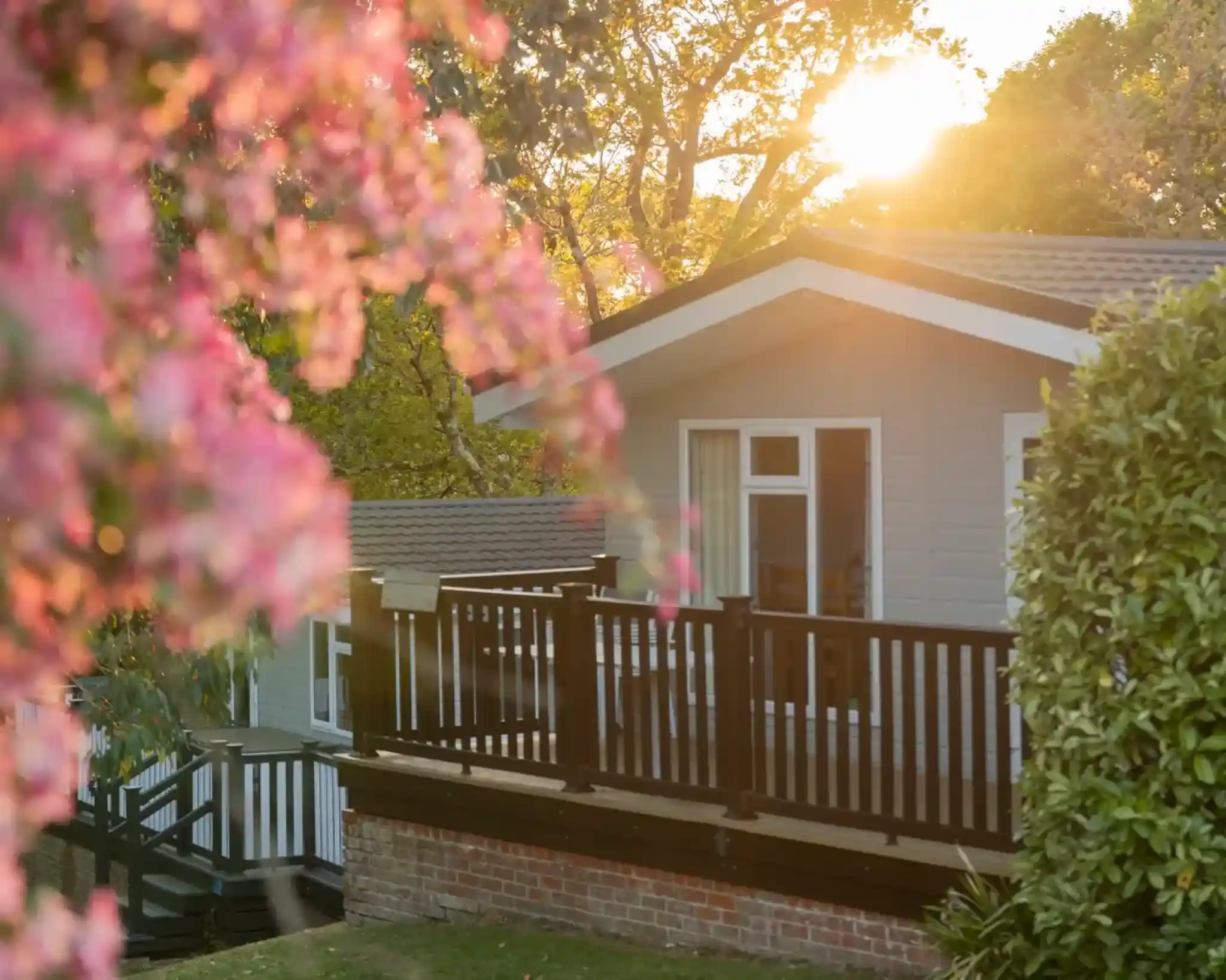 A cozy house with a wooden deck surrounded by lush greenery and blooming pink flowers. The sun is setting in the background, creating a warm, golden glow.