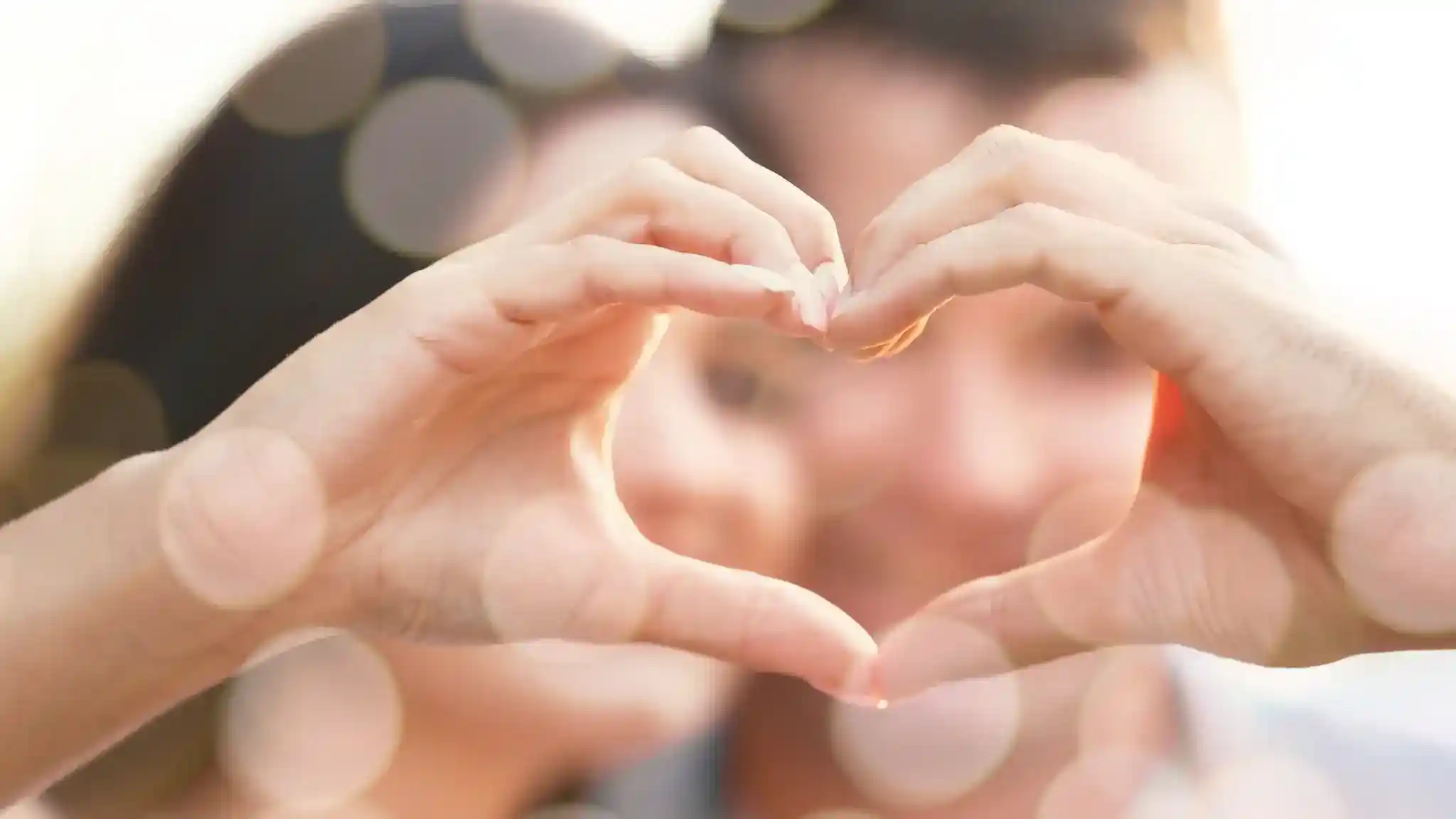 A couple forms a heart shape with their hands while smiling at each other, set against a softly blurred background filled with light bokeh.