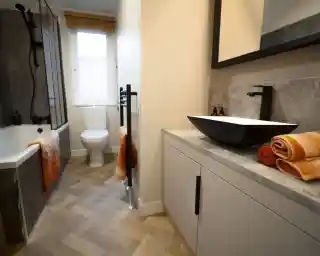 Modern bathroom with a white toilet, bathtub, and a black vessel sink on a light grey vanity, featuring orange towels and a herringbone floor.