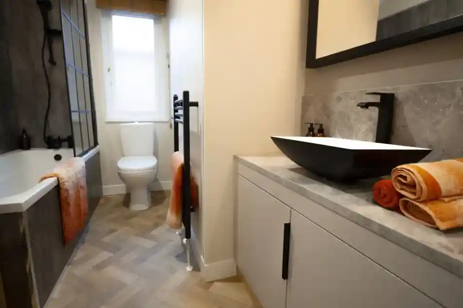 Modern bathroom with a white toilet, bathtub, and a black vessel sink on a light grey vanity, featuring orange towels and a herringbone floor.