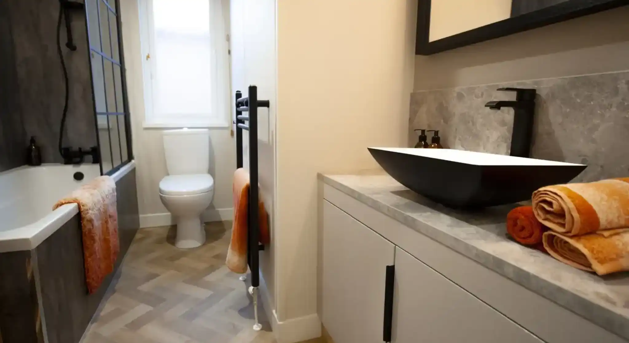 Modern bathroom with a white toilet, bathtub, and a black vessel sink on a light grey vanity, featuring orange towels and a herringbone floor.