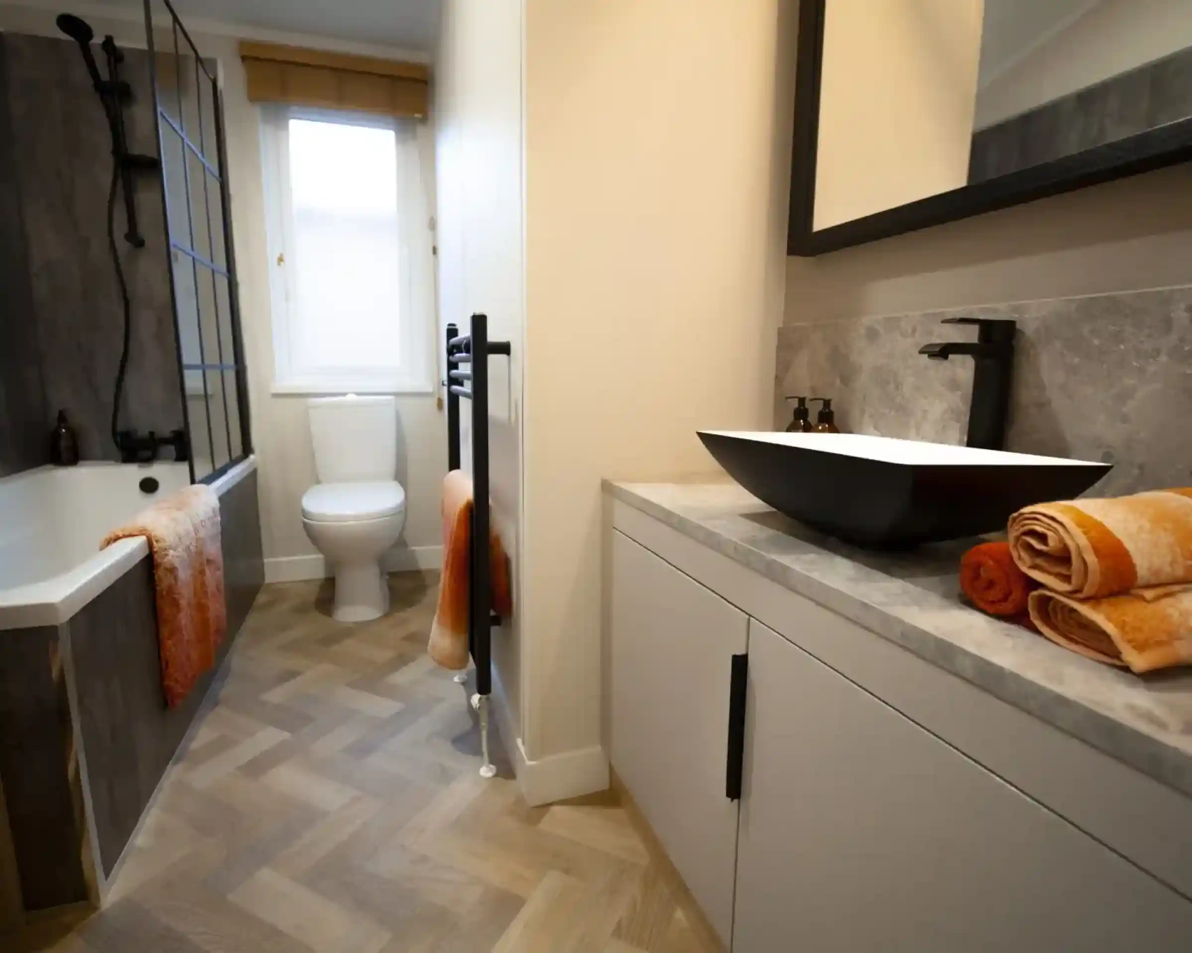 Modern bathroom with a white toilet, bathtub, and a black vessel sink on a light grey vanity, featuring orange towels and a herringbone floor.