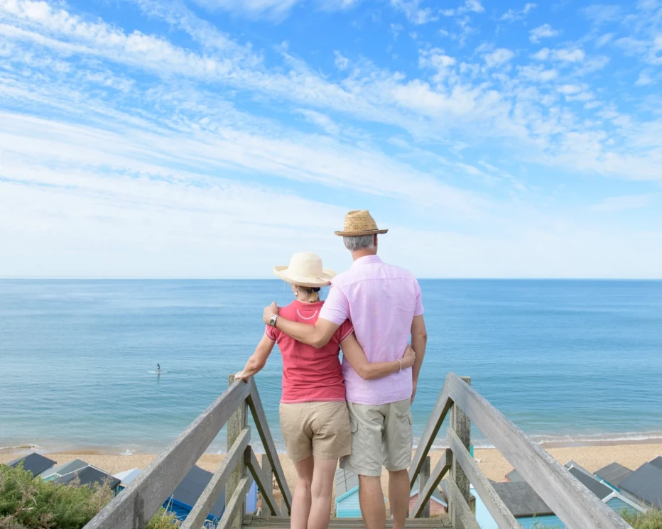 A couple stands on a wooden staircase overlooking a calm sea, with beach huts in the foreground. The sky is bright with scattered clouds. The woman wears a wide-brimmed hat and a red shirt, while the man has a straw hat and a light pink shirt. They are embracing and gazing at the water.