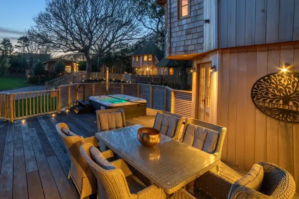 A cozy outdoor deck features a large dining table with wicker chairs and a hot tub. The area is illuminated with soft lighting, surrounded by trees and charming buildings in the background at dusk.