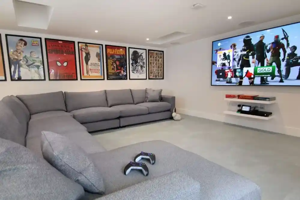 A cozy entertainment room featuring a large gray sectional sofa, two game controllers on the couch, and a big flat-screen TV mounted on the wall. The walls are adorned with various movie posters, creating a vibrant atmosphere.