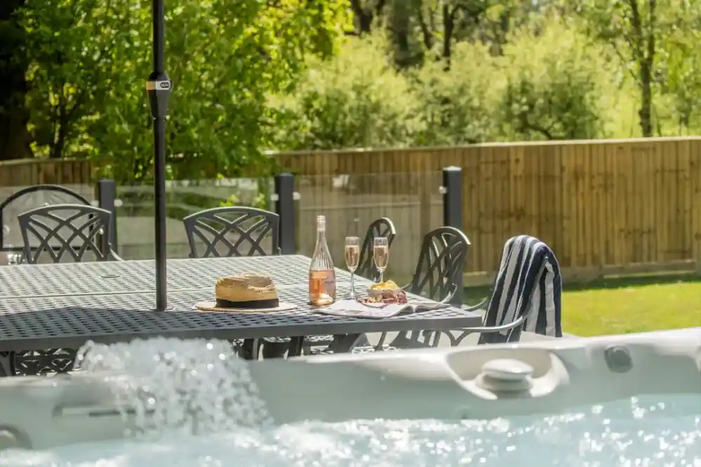 A luxurious outdoor setting features a bubbling hot tub in the foreground, with a dining table and chairs nearby. On the table, there is a bottle of rosé wine, two flutes filled with sparkling wine, a straw hat, and a small plate of snacks. Lush greenery and a wooden fence serve as the backdrop, creating a serene atmosphere.