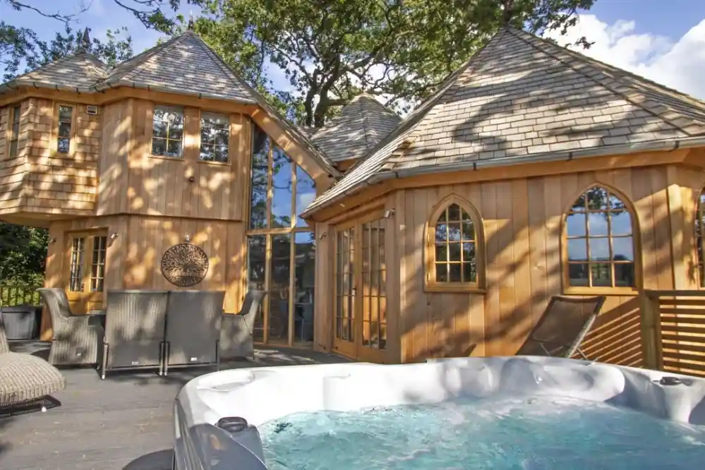 A cozy wooden house with unique architecture, featuring arched windows and a peaked roof. In the foreground, a hot tub bubbles on a sunlit deck surrounded by trees.