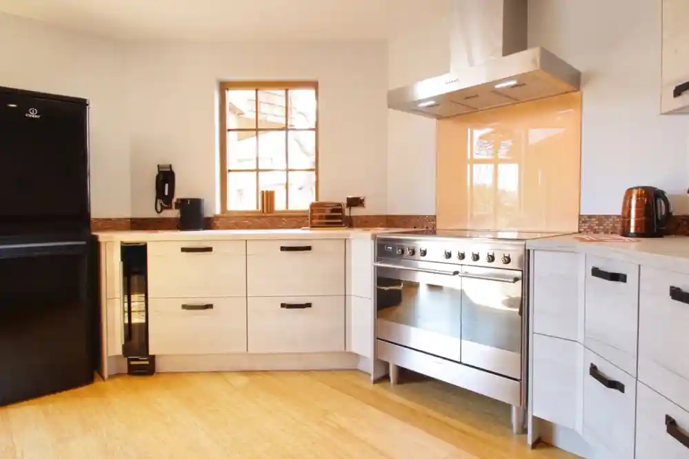 Modern kitchen featuring a black refrigerator, stainless steel oven, and a kettle on the counter. Cabinets are light wood with minimalist black handles, and a large window lets in natural light.