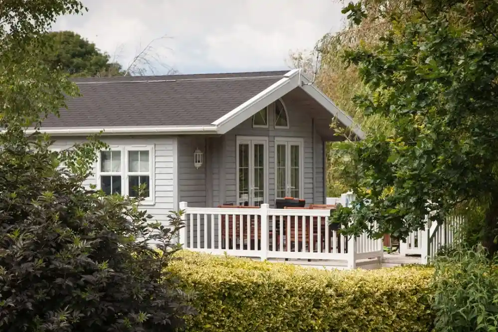 A cozy gray cottage with a porch, surrounded by lush greenery and a low hedge. The house features large windows and a gabled roof.
