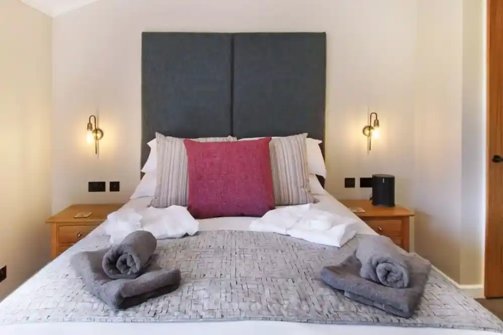 A neatly made bed with a large grey headboard, accented by a pink decorative pillow and striped pillows. Two rolled towels in grey and white are arranged at the foot of the bed, along with a grey blanket. Soft lighting from wall sconces creates a cozy atmosphere.