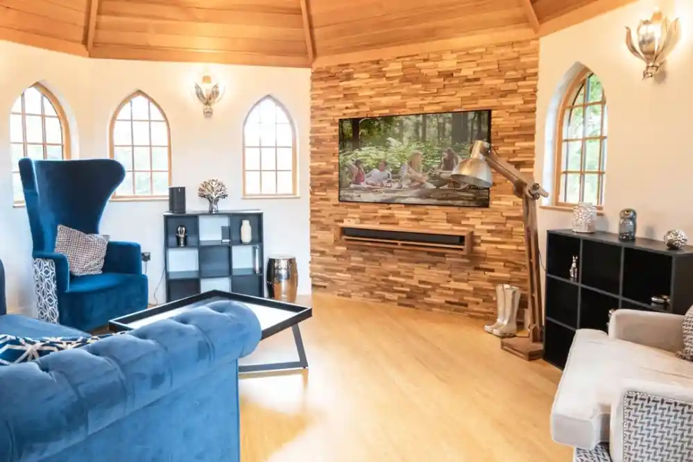 Modern living room featuring a blue wingback chair and a light gray sofa. A large flat-screen TV is mounted on a textured stone wall. The room has wooden ceilings and large arched windows. Stylish decor elements include a black bookshelf and a metallic floor lamp.