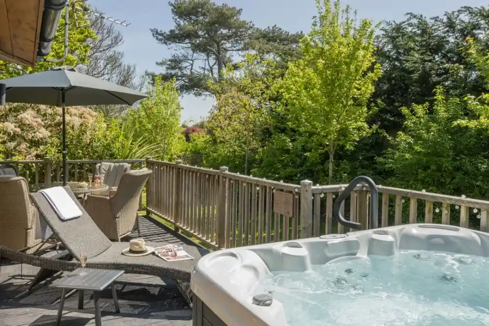 A tranquil outdoor deck featuring a hot tub, two cushioned chairs, and a small table with magazines. Lush greenery surrounds the area, with a clear blue sky above. An umbrella provides shade over the seating.