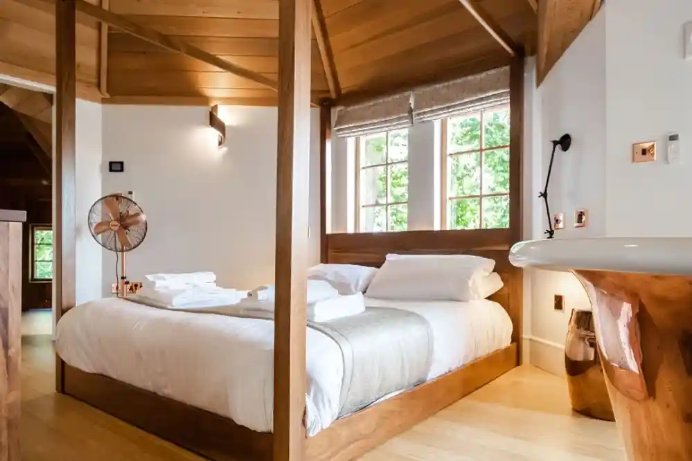 A cozy bedroom featuring a wooden bed with white linens, neatly arranged towels, and large windows providing natural light. The room has a warm wooden ceiling and modern decor, including a decorative fan and stylish light fixtures.