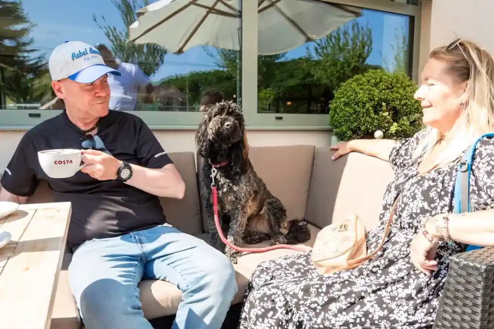 A man in a baseball cap sits on a patio, sipping coffee, alongside a woman in a floral dress. A black dog sits between them, enjoying the sunny outdoor setting with greenery in the background.