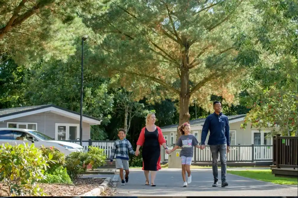 A woman and two children, a boy and a girl, walk hand in hand with a man along a pathway in a green area, surrounded by trees and vacation-style cabins.