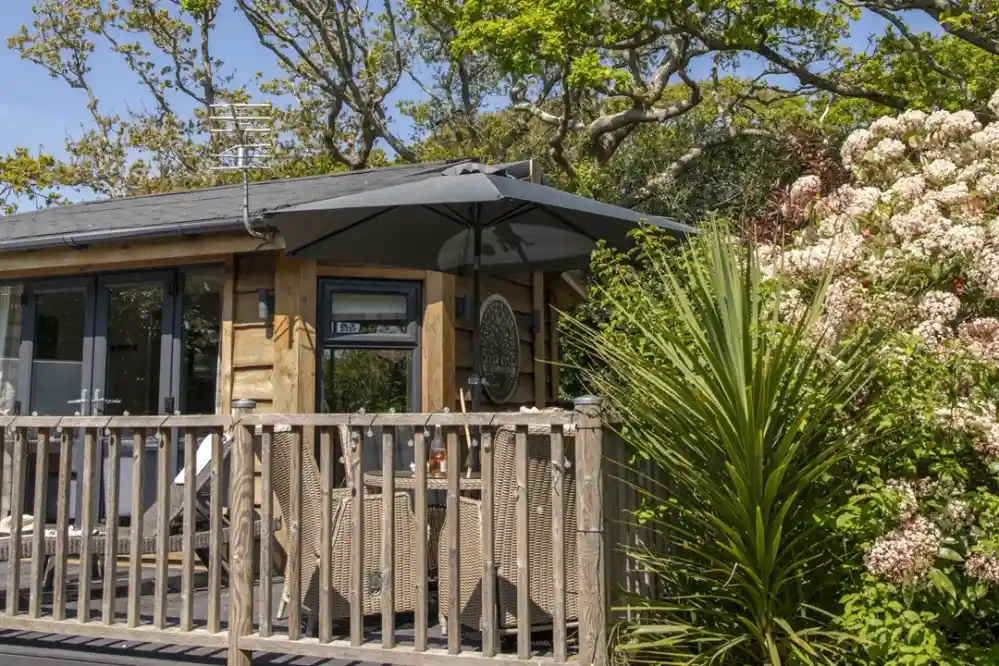 A cozy wooden cabin with a deck, featuring a large umbrella and outdoor seating. Lush greenery and blooming flowers surround the cabin, adding to the serene atmosphere.