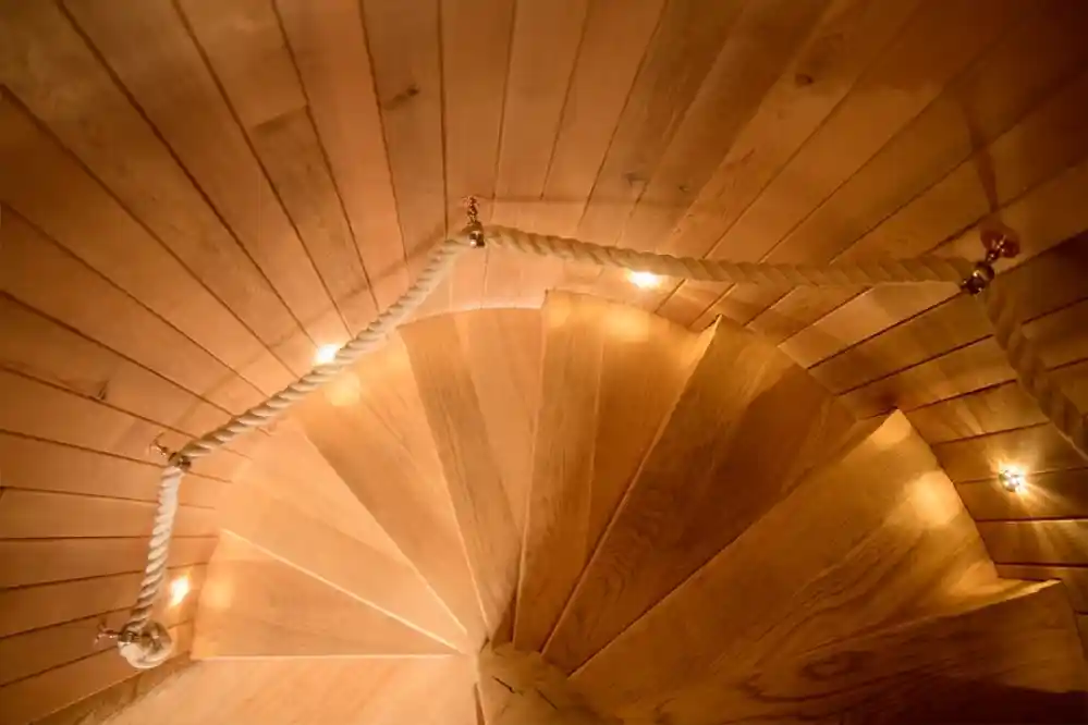 A spiral wooden staircase viewed from above, featuring soft lighting and a rope railing.