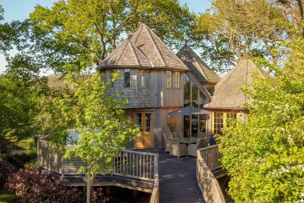 A charming wooden house with a unique design featuring multiple pointed roofs and large windows. Surrounded by lush greenery, a wooden deck with outdoor seating overlooks the landscape.
