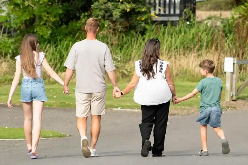 A family of four walks hand-in-hand down a path, with two children on the left and a man and woman on the right. They appear relaxed and are enjoying a sunny day outdoors.