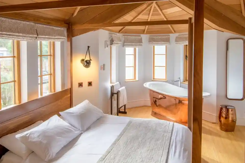 A cozy bedroom with a wooden bed frame and white linens, featuring large windows that let in natural light. There's a freestanding copper bathtub and a modern bathroom setup, along with a warm wooden aesthetic throughout the space.