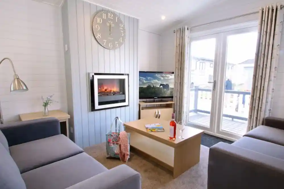 A cozy living room featuring two gray sofas, a wooden coffee table, and a flat-screen TV. A modern electric fireplace is mounted on the wall under a large clock. Large glass doors lead to an outdoor balcony. A floral vase and a backpack with a drink are placed on the coffee table.