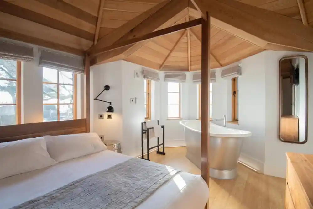 A modern bedroom featuring a circular wooden ceiling and large windows. The bed is neatly made with a light gray throw. In the corner, there is a sleek, freestanding bathtub next to a small stand. A mirror and a stylish light fixture add to the contemporary decor.
